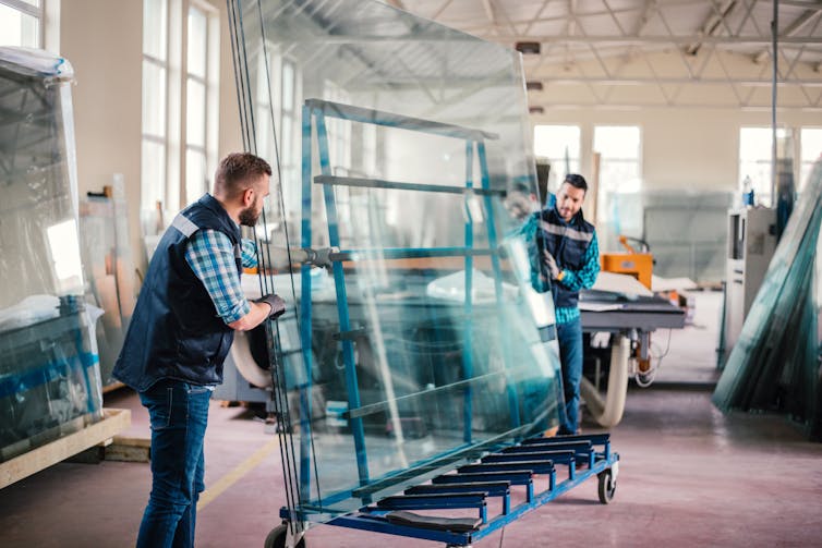Two men wearing gloves roll large sheets of clear glass, taller than themselves, on a cart.
