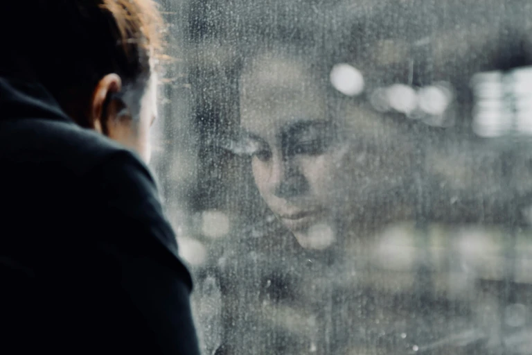 The reflection of a contemplative young woman in a window covered in raindrops