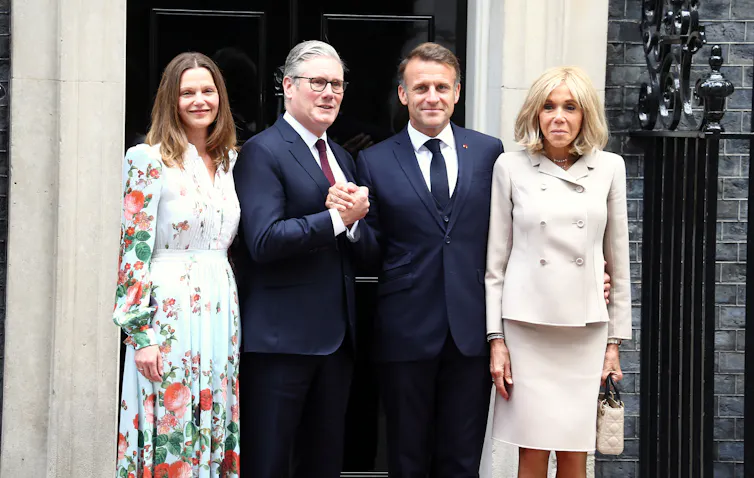 Two middle-aged couples, men in dark suits, women in summer wear standing on the steps of 10 Downing Street in London.