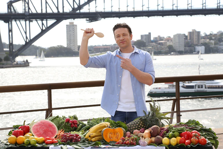 Jamie Oliver holding a wooden spoon. He's standing behind a table covered in various fruit and vegetables.