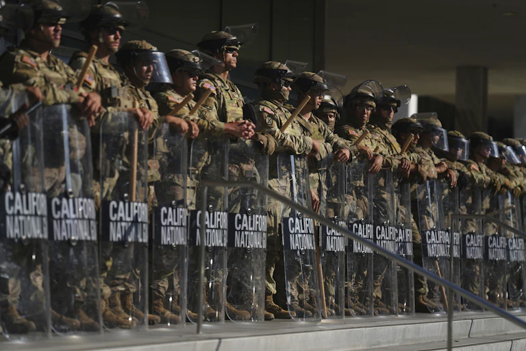 Several soldiers wearing helmets and holding batons and shields stand in a row.