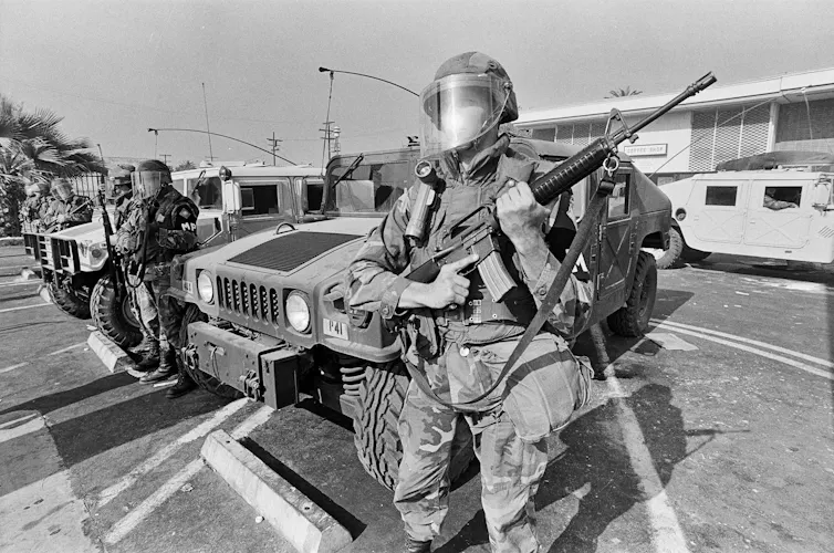 Soldiers holding machine guns and grenade launchers stand on a street in Los Angeles.