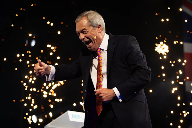 Nigel Farage gesturing to the crowd while giving a speech. Indoor fireworks go off in the background.