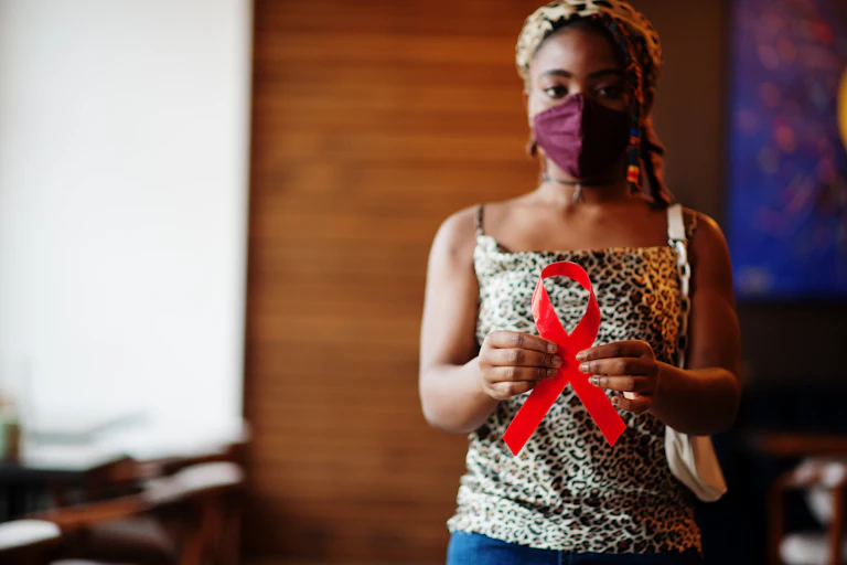 Une jeune femme, avec un masque chirurgical sur le visage, tient dans la main le ruban rouge symbole de la lutte contre le VIH.