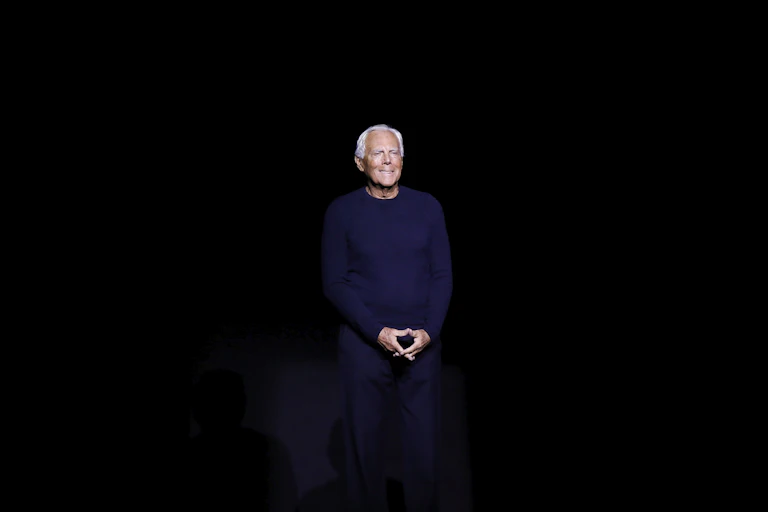 A smiling white-haired man stands on a darkened stage.