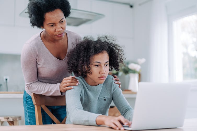 a Black woman stands behind a black teen girl seated at a table using a laptop