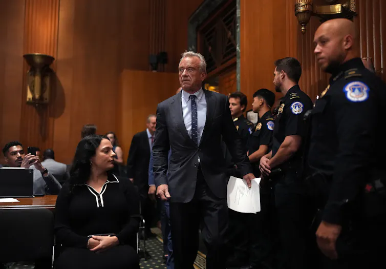 HHS Secretary RFK Jr. walks into a Senate office building, flanked by police officers standing along the wall.