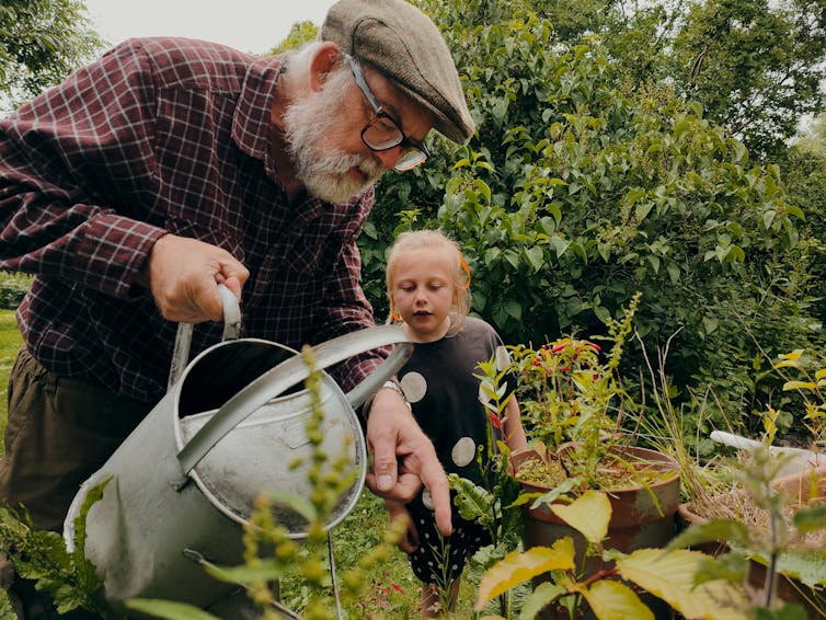 An old man with a watering can points to a plant for a young girl