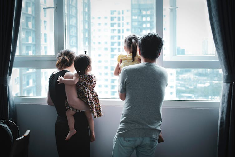 The backs of a man and a woman, each holding a child, looking out a window at high-rise apartment buildings.