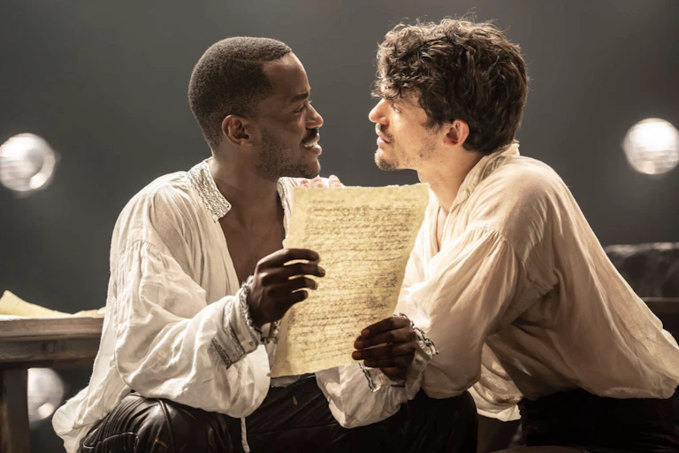Two men sitting very close together staring into each other's eyes, one holding a piece of paper with writing on it.
