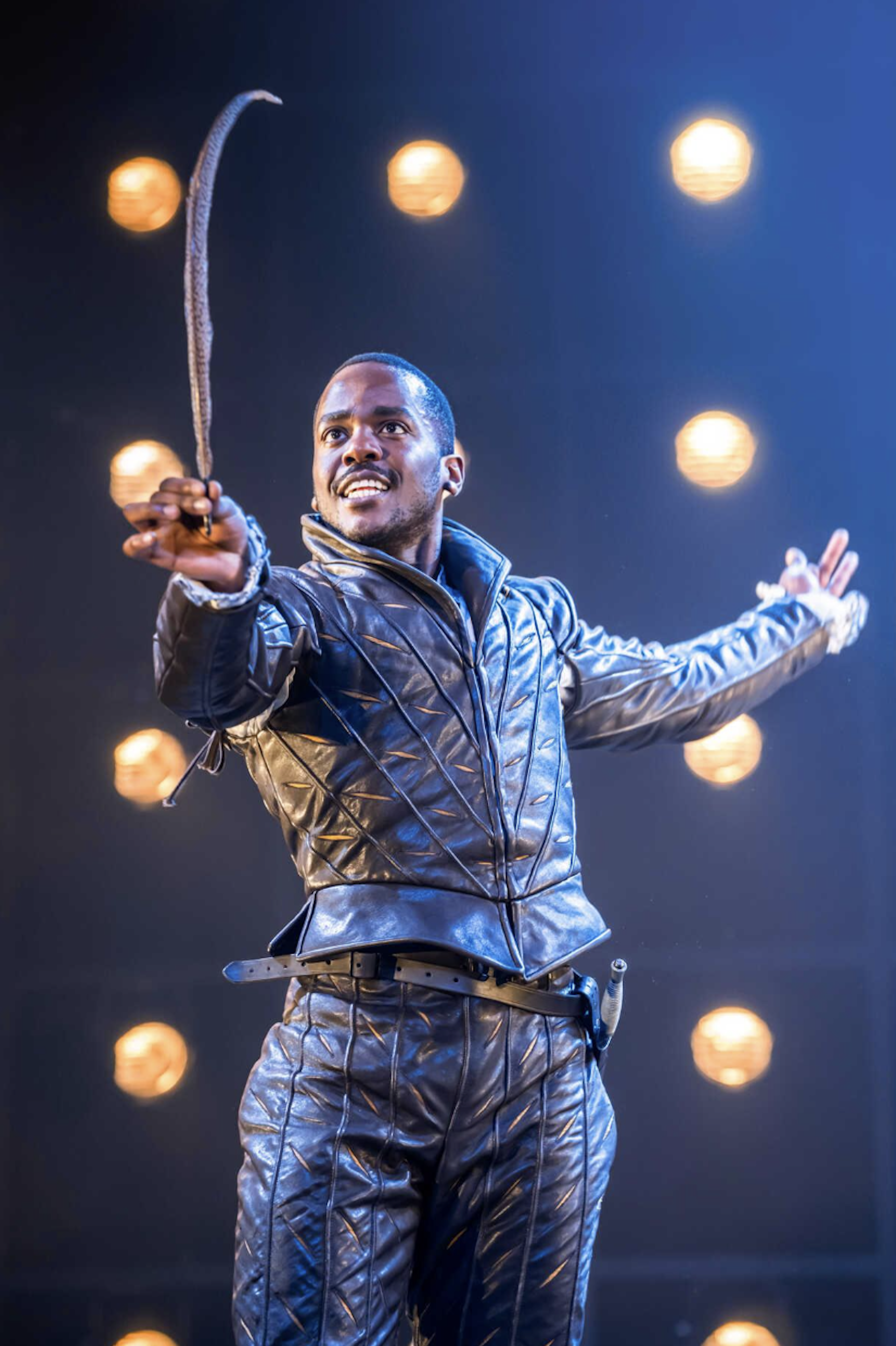 An actor on stage in Elizabethan costume holding a feather quill in his hand.