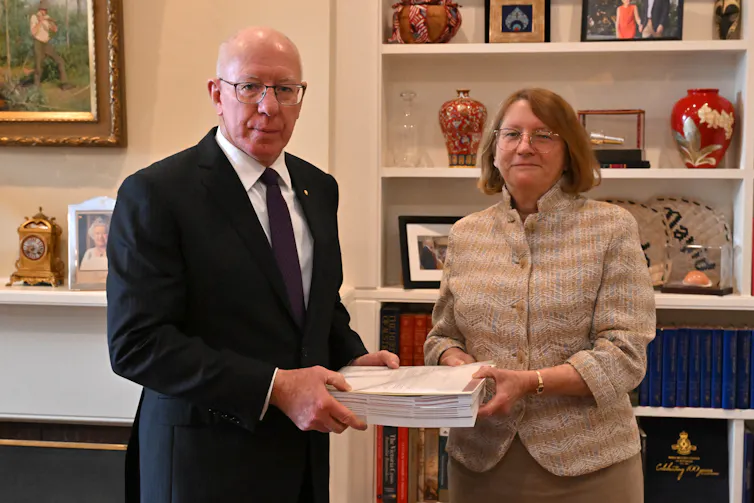 A man and a woman look at the camera while holding a large stack of papers