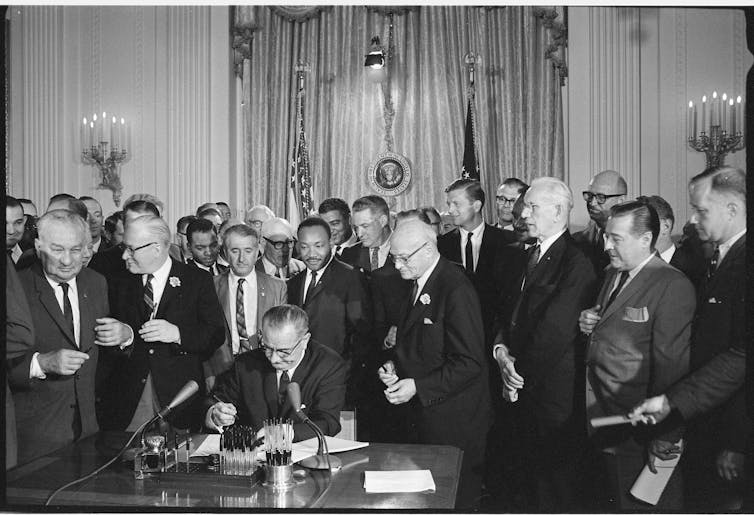 A black and white photo of President Lyndon B. Johnson signing a document surrounded by many men.