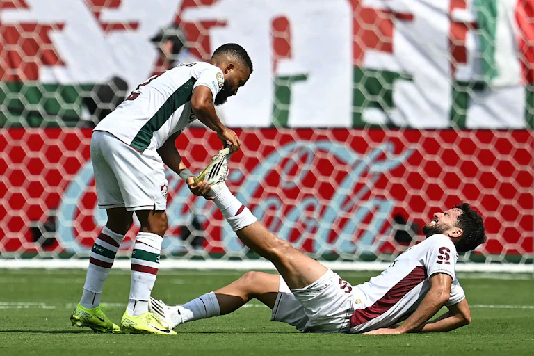 A Fluminense soccer player stretching the cramped leg of a teammate lying on the ground, their faces held in a grimace.