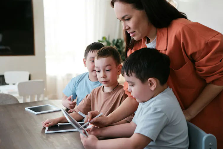 A woman watching over kids who are using technological devices at home.
