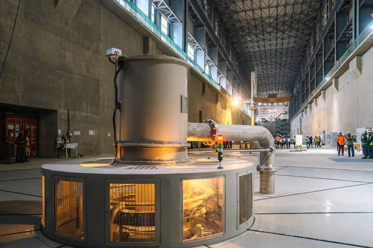 A large steel turbine in the foreground is lit up while groups of people stand off to the side in the background