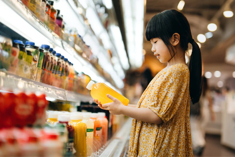 Young girl looks at juice bottle in sweetened drinks aisle.