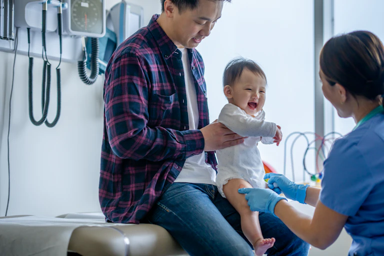 A father holds a small child who is seen by a nurse.