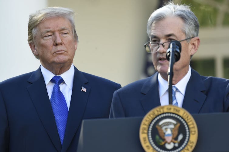 Two men in dark blue suits, one standing behind a lectern and microphone.