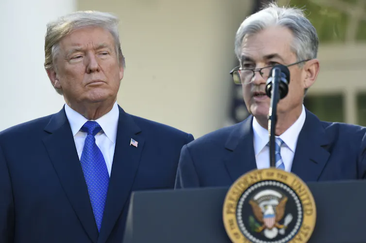 Two men in dark blue suits, one standing behind a lectern and microphone.