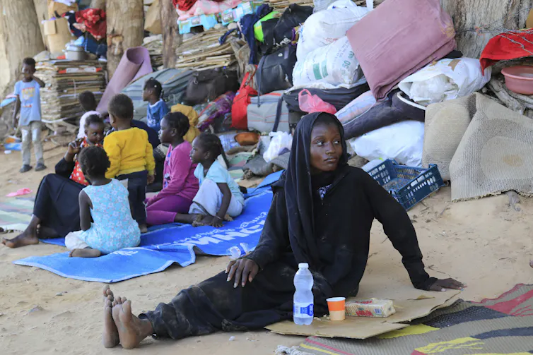 A Black woman sits on the ground among other refugees gathered nearby.
