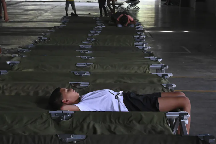 A young boy in a white T-shirt lies on his back on a cot looking up at the ceiling.