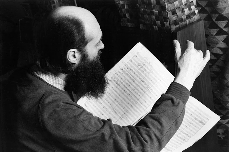 A black and white photograph of a balding man, seen in profile, who holds a musical score and holds up one hand as though conducting.