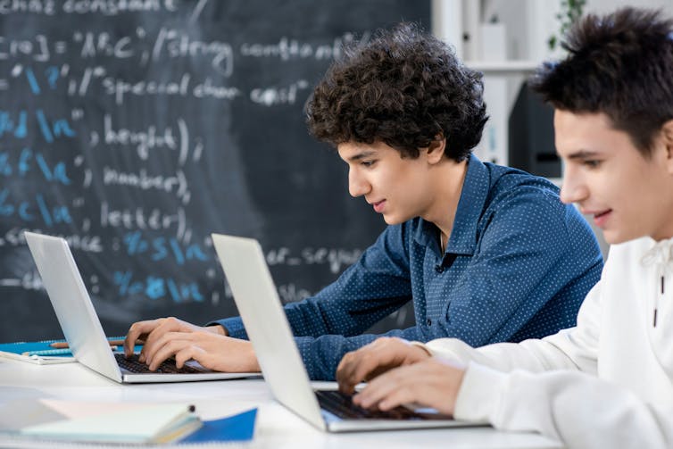 two students sitting at laptops with equations on a blackboard behind them
