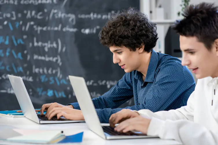 two students sitting at laptops with equations on a blackboard behind them