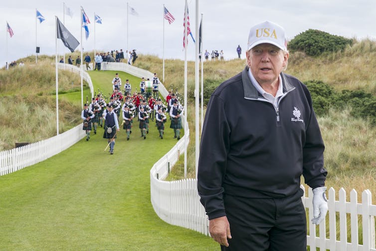 A man in a white cap and golf glove stands on a patch of grass with bagpipers behind him