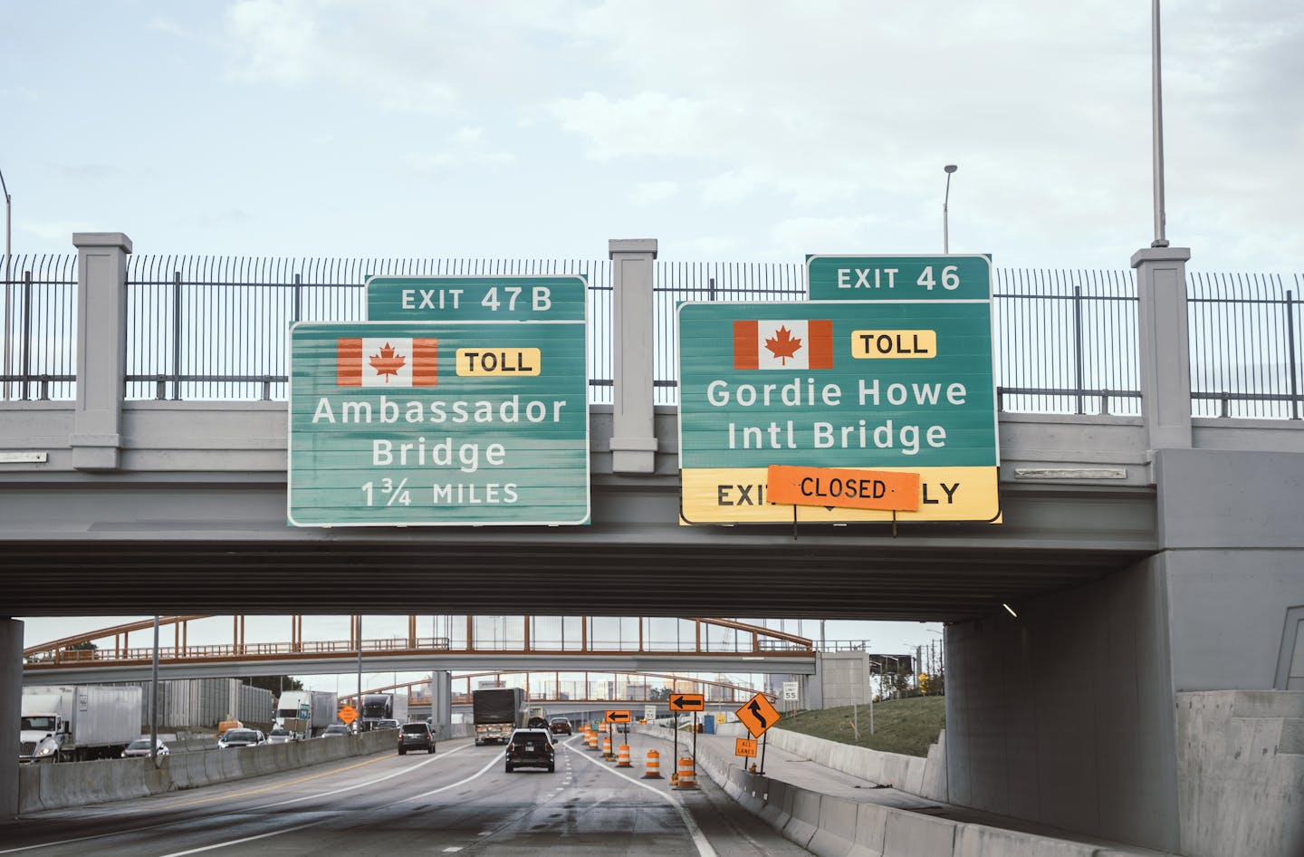 Large signs affixed to a bridge over a highway, in white lettering on green signs, show the exits for the Ambassador Bridge and the closed Gordie Howe International Bridge.