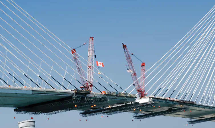 Canadian and American flags are held by cranes on either side of a large suspension bridge.