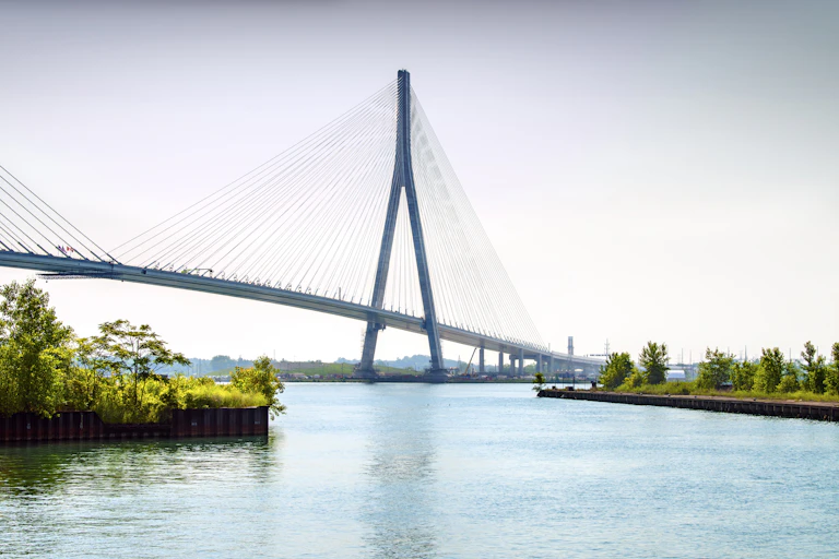 A large cable bridge spans a large expanse of water. Green marsh is on either side of blue water and a city is barely visible in the distance