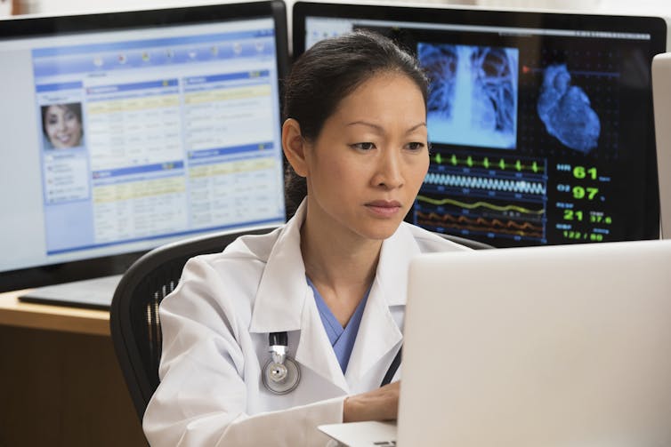 A doctor is flanked by computer screens and working on a laptop.