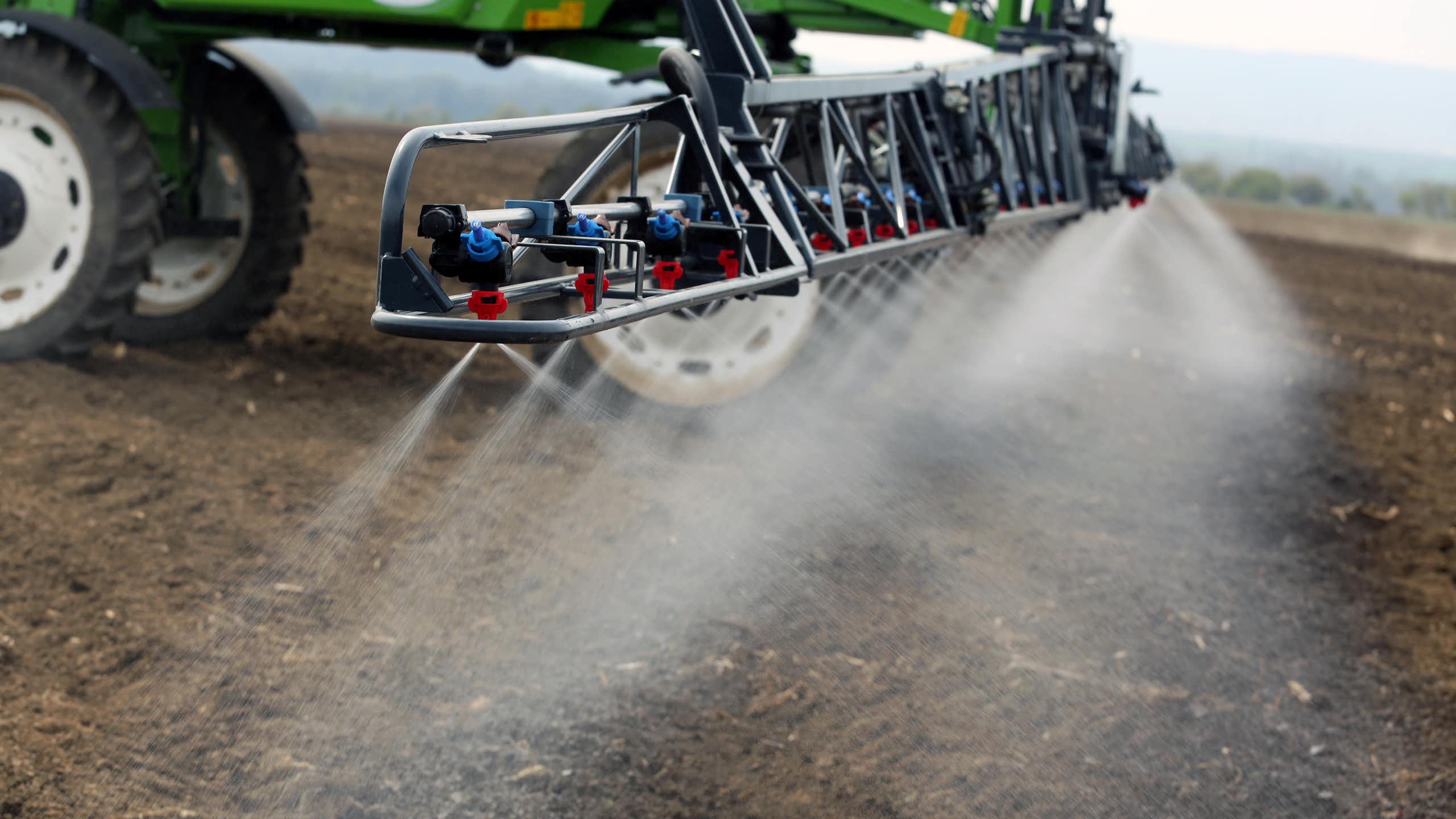 A sprayer at the rear of a tractor sprays a field of land with a white liquid.