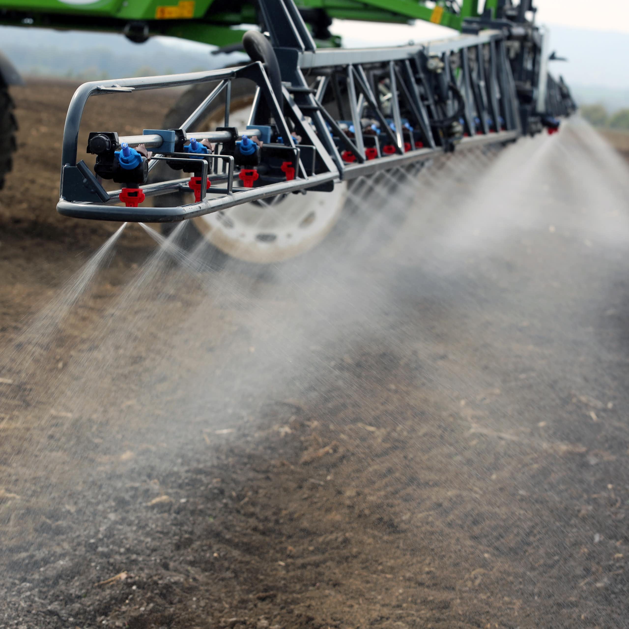 A sprayer at the rear of a tractor sprays a field of land with a white liquid.