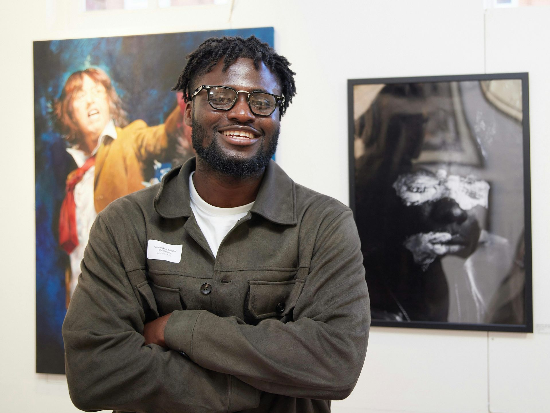 An African man smiles for the camera, arms crossed. Behind him are framed artworks on a wall.
