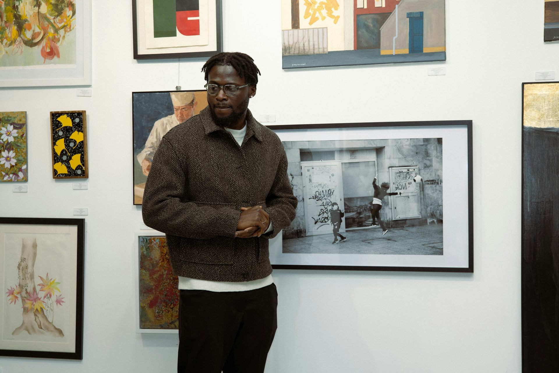 A young African man looks to one side as he stands in front of a wall of paintings and photos.