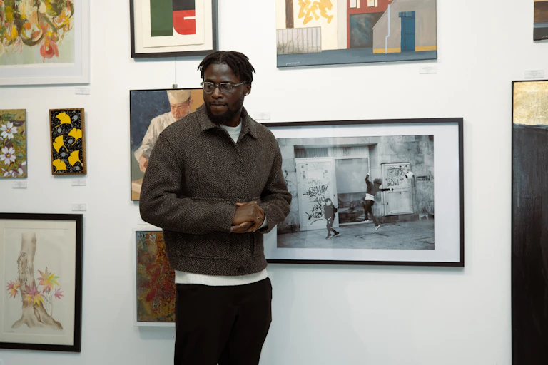 A young African man looks to one side as he stands in front of a wall of paintings and photos.