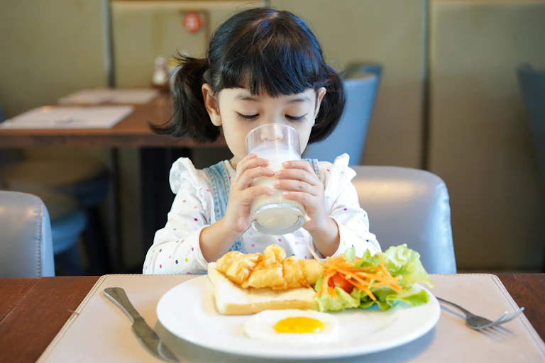 A young girl drinks a glass of milk. She has a plate of food in front of her with chicken, salad and a fried egg.