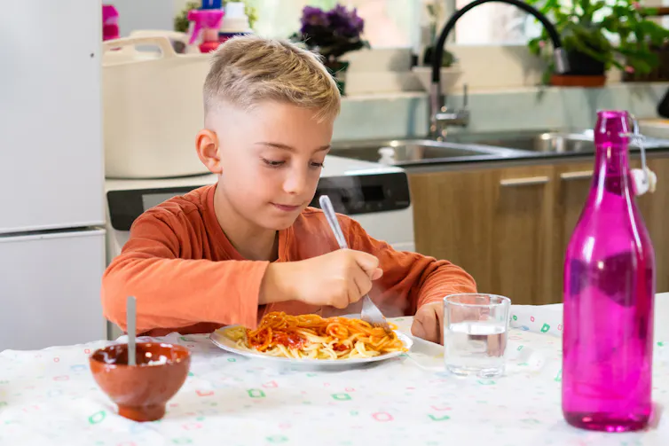 A boy uses a fork to eat a plate of spaghetti with red sauce.