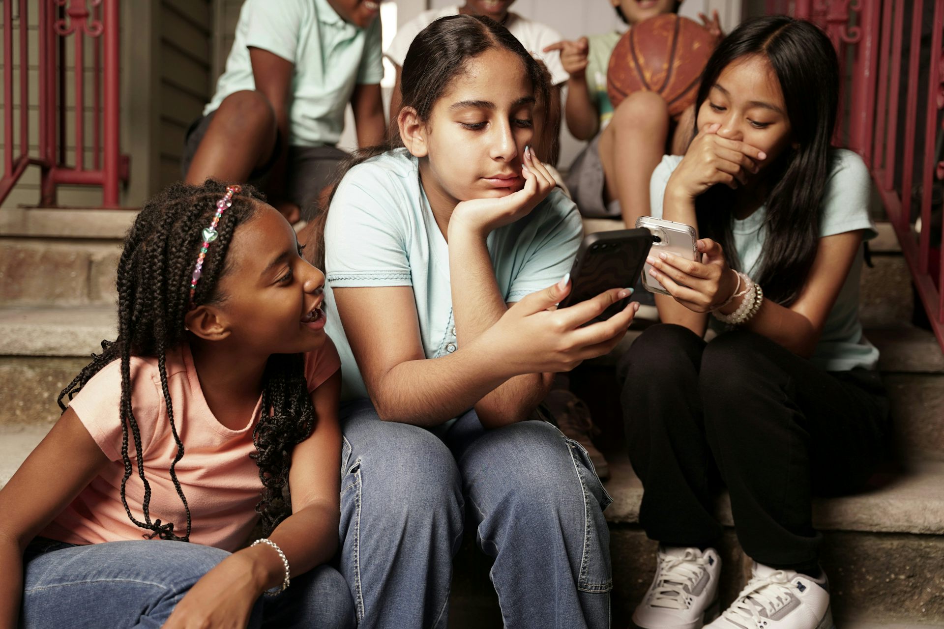 Three girls at school are huddled together on stairs looking at two cellphones.