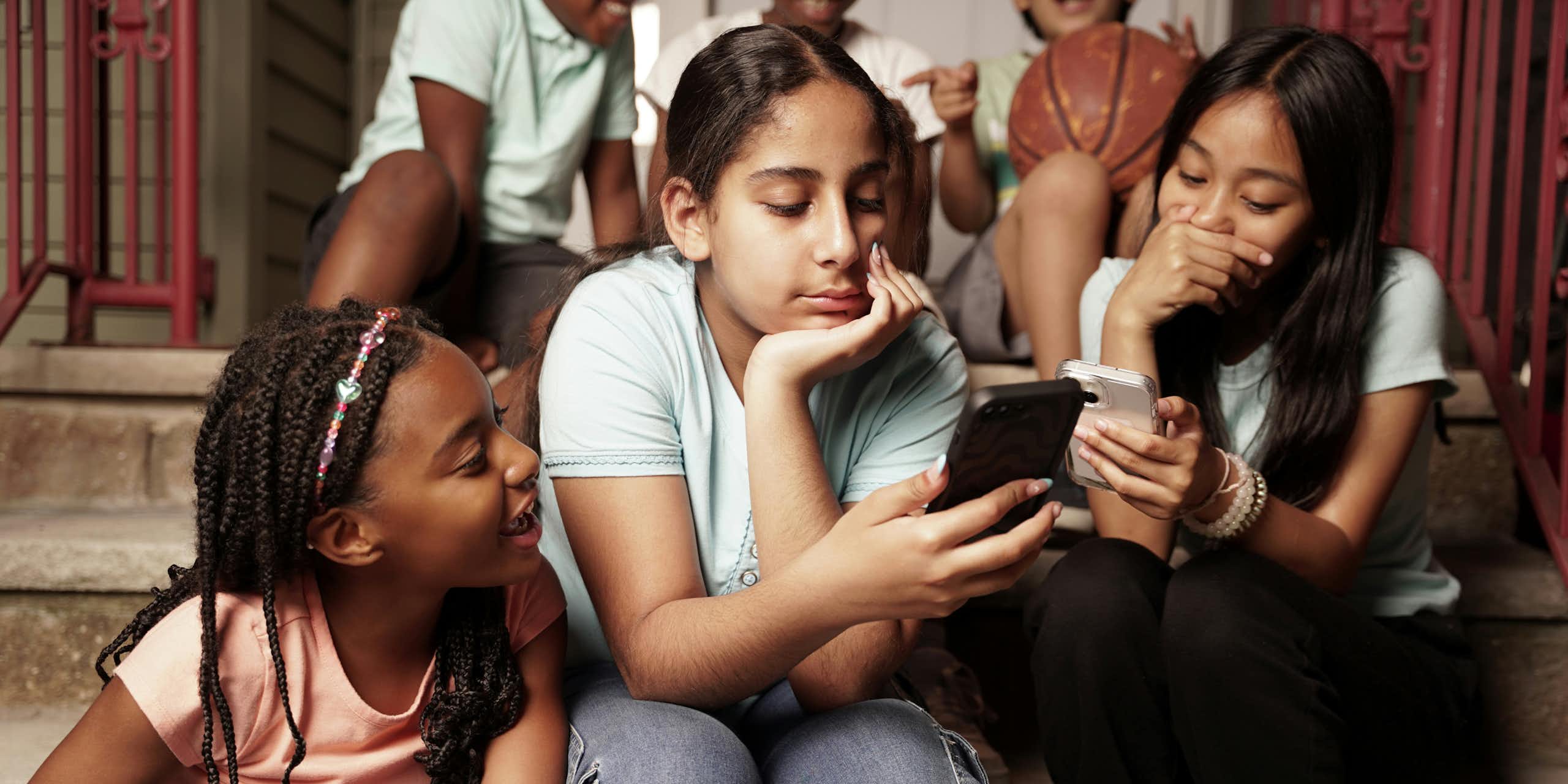Three girls at school are huddled together on stairs looking at two cellphones.