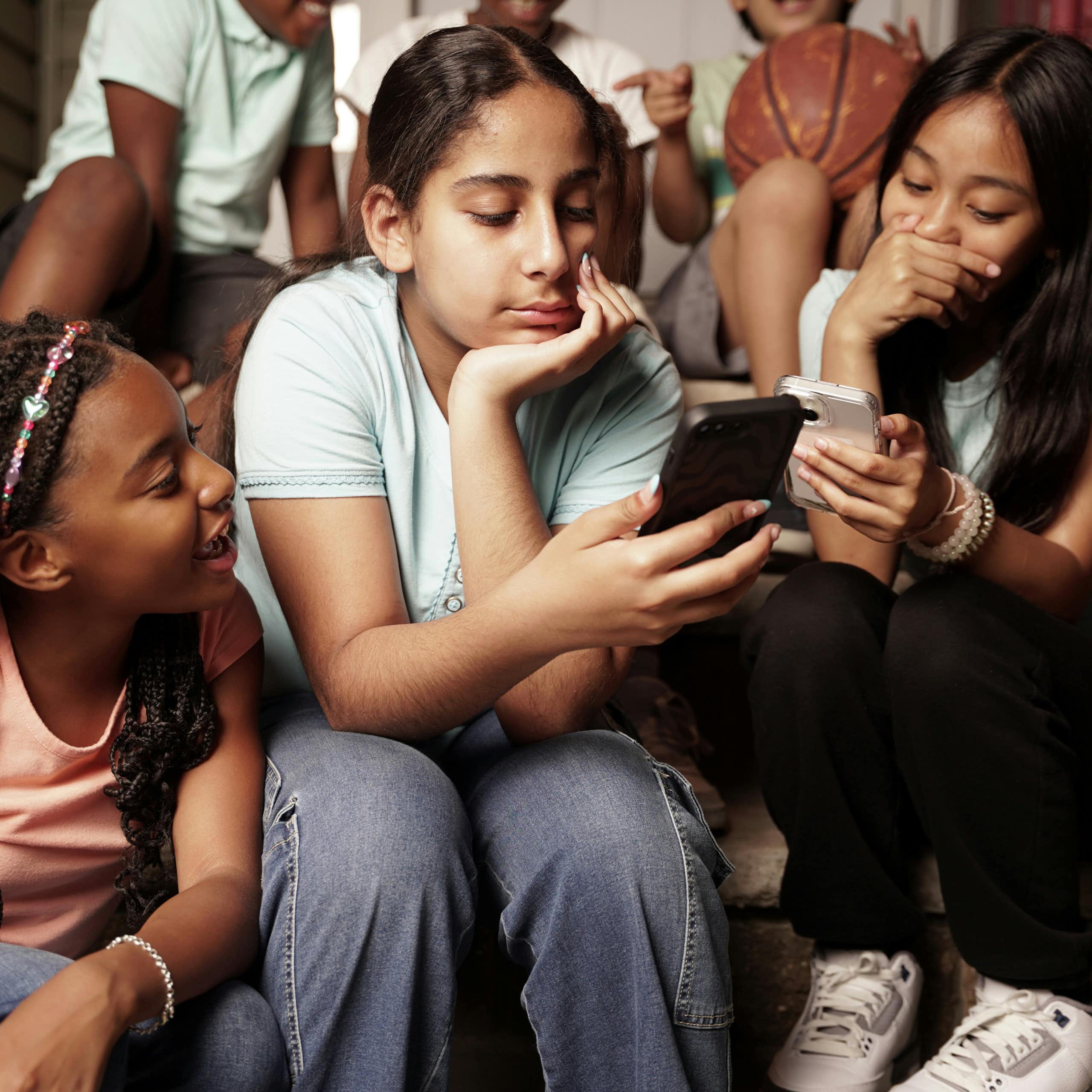 Three girls at school are huddled together on stairs looking at two cellphones.