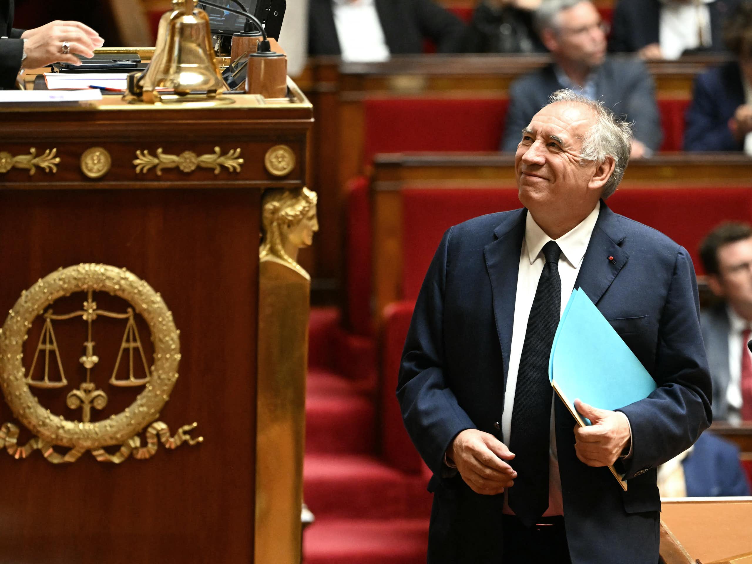 François Bayrou, the prime minister of France, is seen standing and holding a folder in the National Assembly.