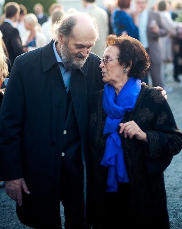 An older man in a dark outfit and a woman in a bright blue scarf lean against each other as they talk and walk outside.