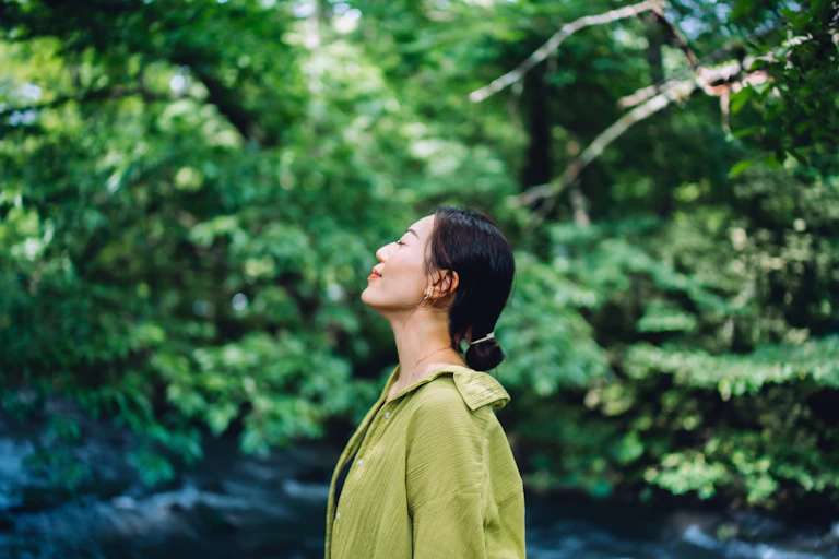 Young woman standing with her eyes closed, appearing to meditate, with lush trees in the background.