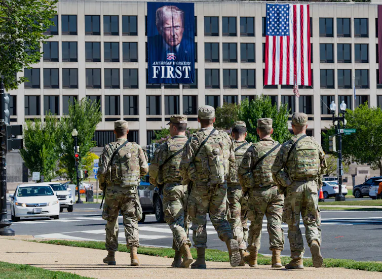 A group of armed soldiers walk in front of a building on which hangs a large banner of Donald Trump.