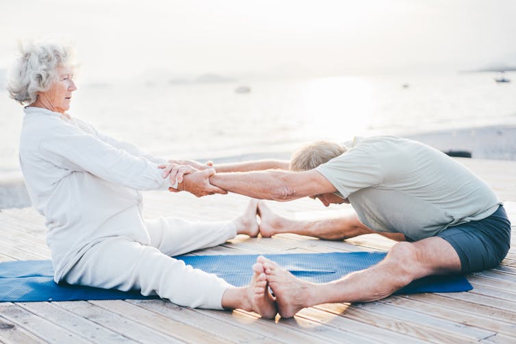 A senior couple sitting on the beach, pressing their feet together as the woman pulls the man's arms forward in a stretch.