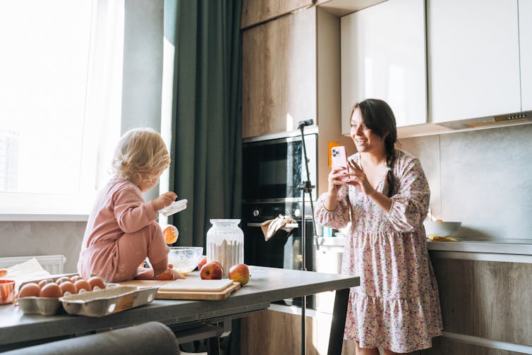 A woman in a pink dress holds her phone in a kitchen as she films a young child sitting on a countertop besides a carton of eggs.